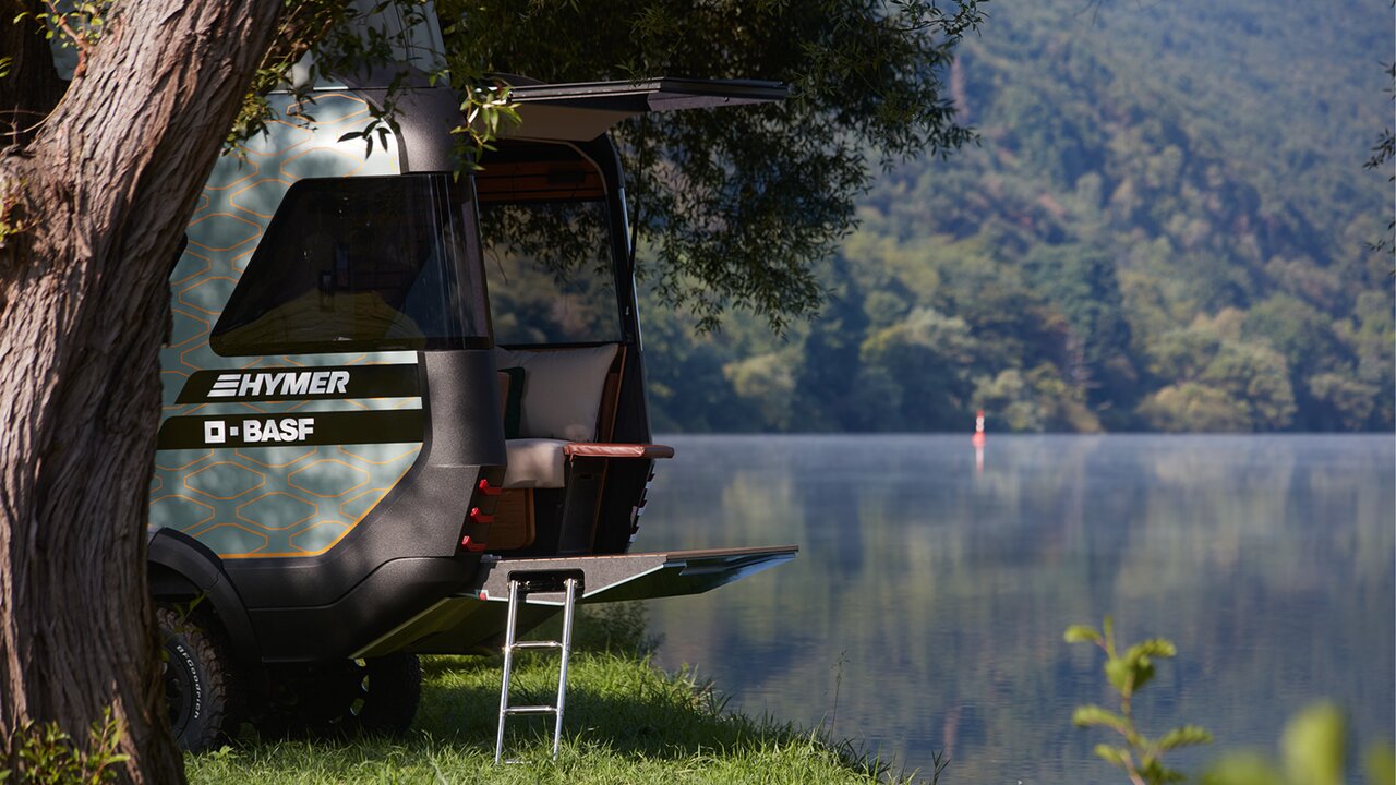 The HYMER VisionVenture concept vehicle stands on a lake behind a tree in the grass with the rear open and the ladder folded out.