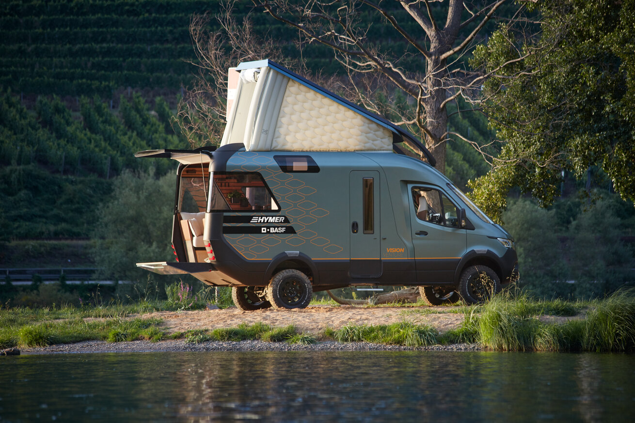 The VisionVenture, a HYMER motorhome, is parked on the shore of a lake with hills in the background; the sleeping loft and rear are open.