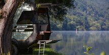 The HYMER VisionVenture concept vehicle stands on a lake behind a tree in the grass with the rear open and the ladder folded out.