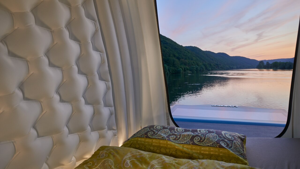 Blick auf Wald und See aus dem geöffneten Schlafbereich des HYMER VisionVenture, der als Loft über dem Mercedes-Wohnmobil liegt. 